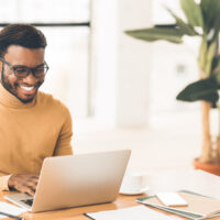 Successful Freelancer. Happy afro man smiling and working on laptop in home office. Copyspace, panorama